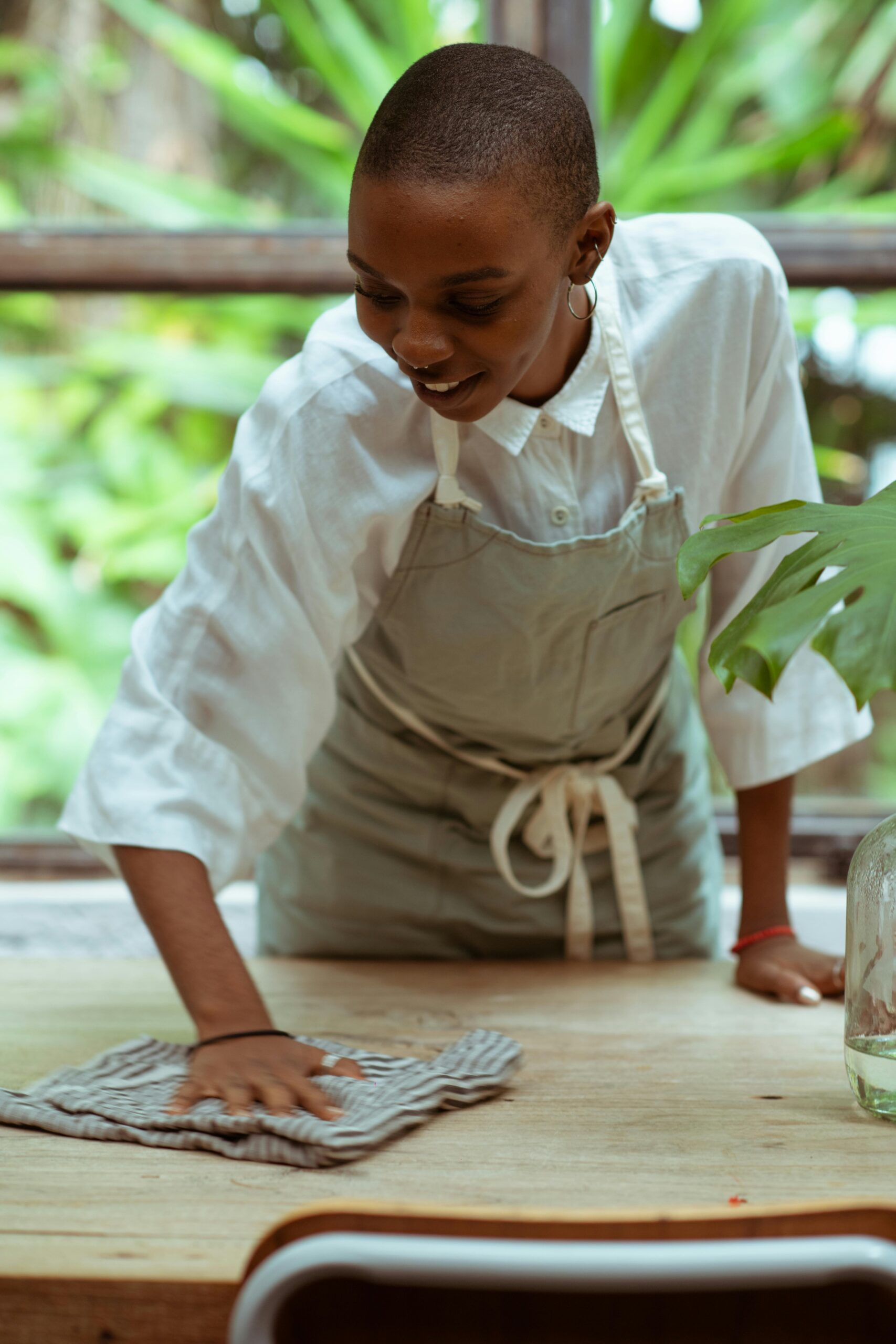 Positive smiling black woman with short haircut wearing apron cleaning wooden table by using microfiber cloth while standing against blurred big window and green lush park